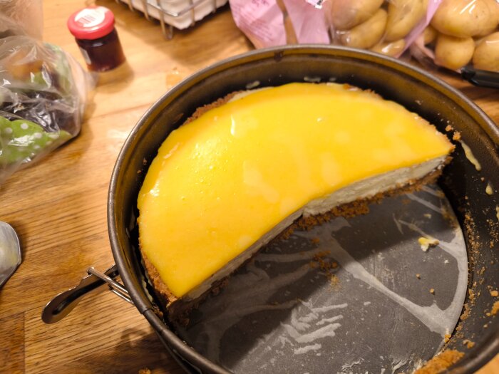 Cheesecake med passionsfrukts curd i en rund bakform på ett köksbord, omgiven av potatis och en burk i bakgrunden.