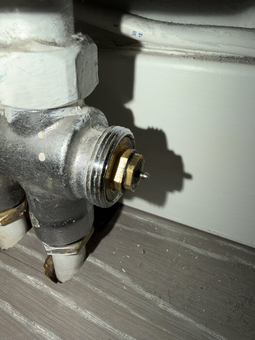 Närbild på en metallventil kopplad till rör, med synlig gänga och mässingsdel, mot en vit vägg och golv med damm.