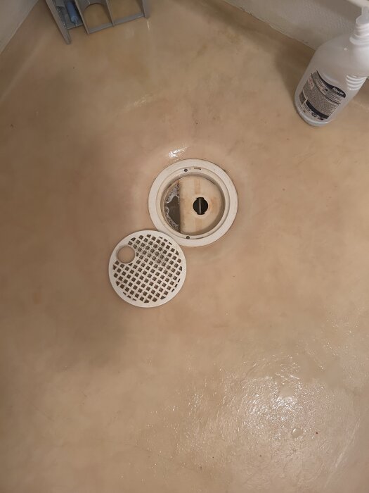Öppen duschbrunn med avtaget galler på ett beige badrumsgolv, bredvid en flaska rengöringsmedel.