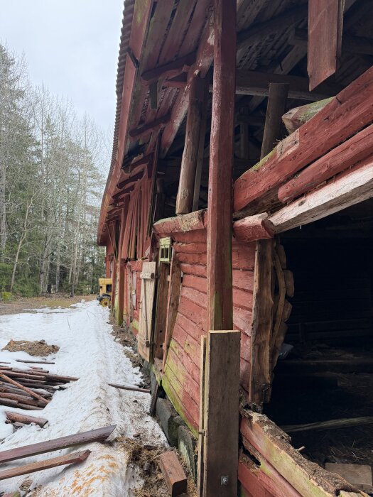 Röd träbyggnad med skadad vägg, flera stolpar synliga, snö på marken, omgiven av träd i bakgrunden.