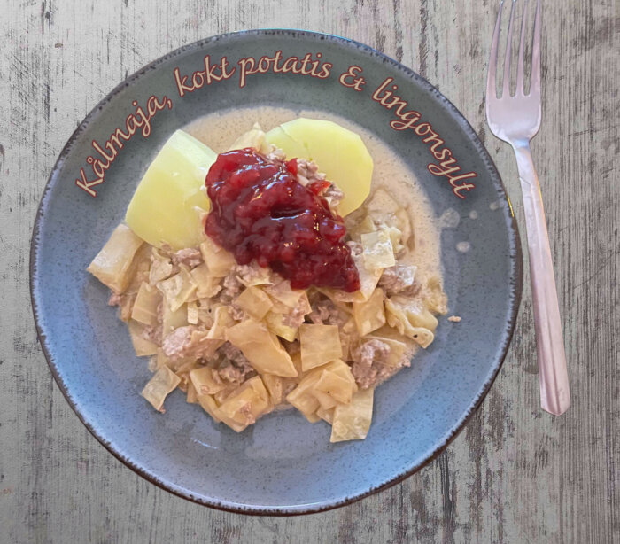 Tallrik med kålpudding, kokt potatis och lingonsylt, med texten "Kålmaja, kokt potatis & lingonsylt" runt kanten.