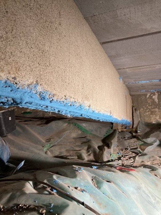 Bilden visar en balk underifrån med blått antikorrosionsskydd och betongreparationer ovan, samt plastskydd på marken under.