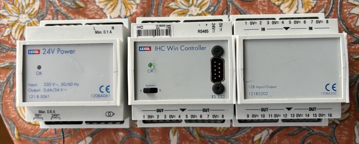 Tre vita elektroniska enheter med etiketter, inklusive en Lexel 24V Power och en IHC Win Controller, placerade på ett blommigt tyg.