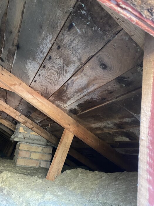 Loft med gamla träbjälkar, synlig isolering och en murad skorsten; tecken på påväxt och smuts.
