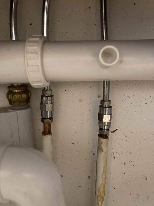 Vattenledningar med rostfläckar och synliga kopplingar under ett handfat.