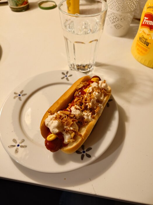 Hot dog med rostad lök och sÄs pÄ en tallrik, bredvid ett glas vatten och en flaska senap pÄ ett bord.