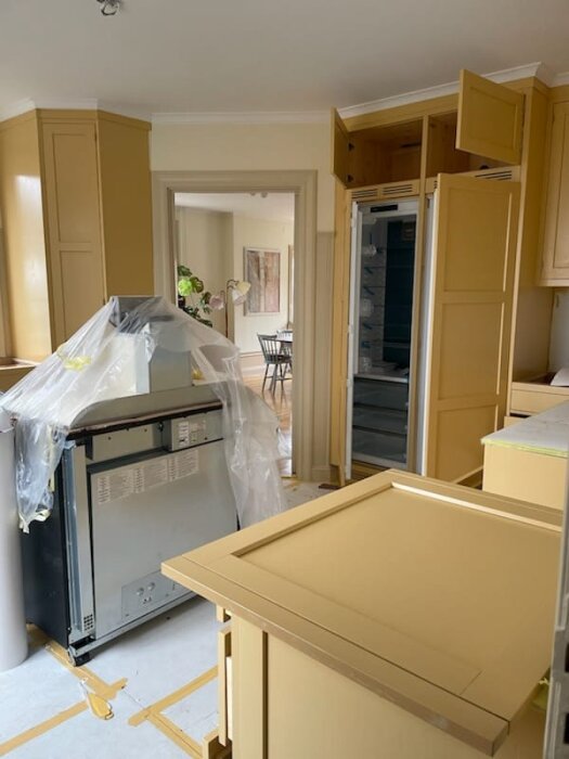 Linoljemålat kök med guldfärgade skåp och plastskyddad köksmaskin i renoveringsprocess.
