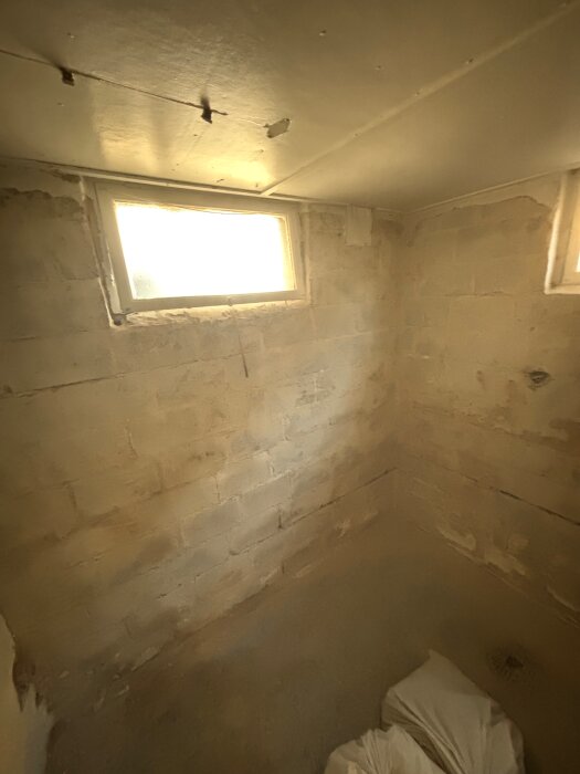 Källarrum under renovering med synliga sandiga väggar, små fönster och oavslutad yta. Ljuset kommer genom ett av fönstren.