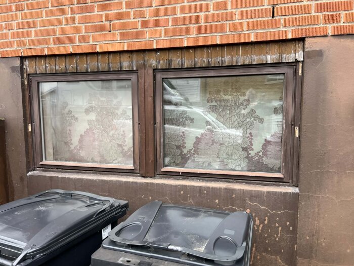 Stort källarfönster med bruna träkarmar i behov av renovering sitter i tegelvägg, två soptunnor syns framför fönstret.