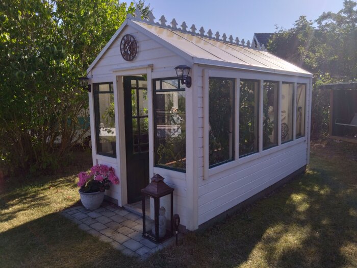 Växthus med vita träväggar och glasrutor, omgivet av grönska. Framför växthuset finns en kruka med rosa blommor och en lykta.