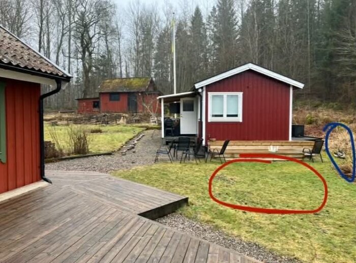 Gräsmatta med rödmarkerad plats för planerad bastu och blåmarkerade tankar nära röda hus samt skog i bakgrunden.