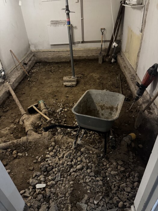 Källare under renovering med grusig jord, en skottkärra, verktyg som spade och borrmaskin samt rördelar på marken.