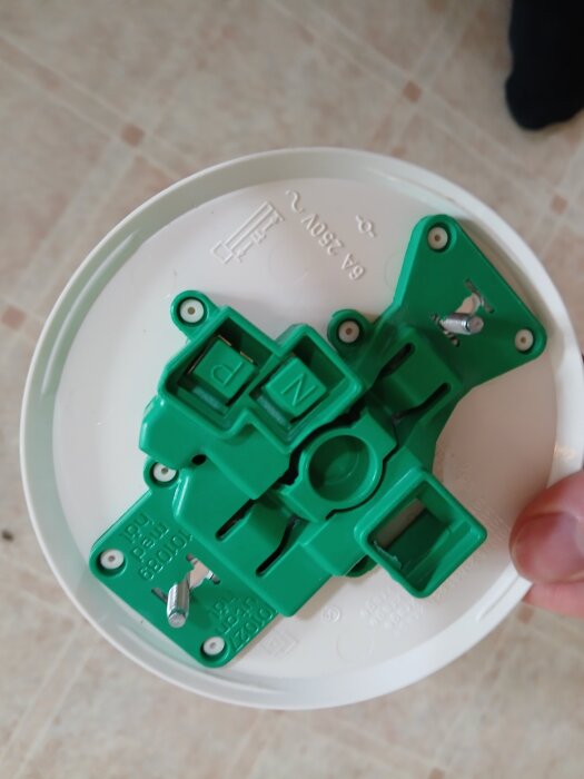 Grön plastdetalj med anslutningspunkter för takkabel på vit platta, text "250V" synlig, hålls av en hand på ljus golvyta.