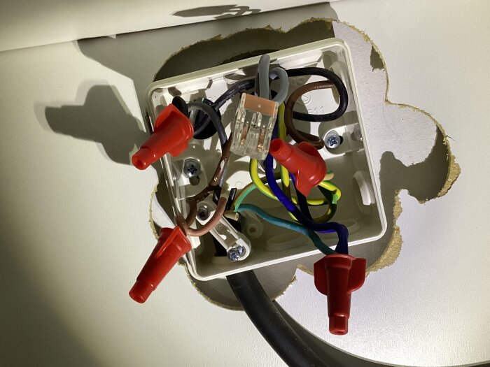 Elektrisk kopplingsdosa med flera kablar i olika färger och röda plastlock, monterad i en vit vägg med ett ovalt hål.