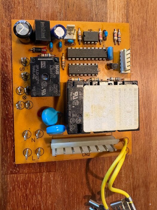 Elektroniskt kretskort med flera komponenter som kondensatorer, motstånd och reläer, liggande på ett träbord.