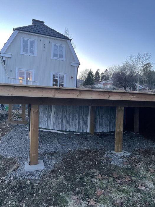 Trästomme till altan under konstruktion framför ett vitt hus med sadeltak. Träpelare står på betongplattor i en grusad yta.