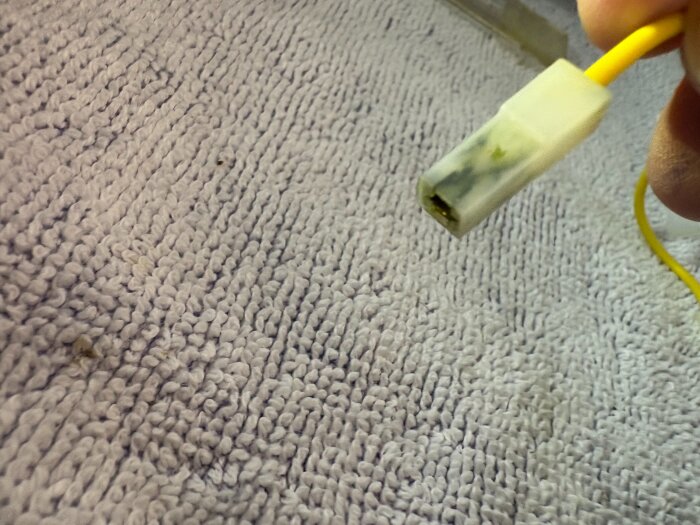 Närbild av en ärgad kabelkontakt med gul sladd, hålls i en hand, mot bakgrund av en texturerad grå yta.