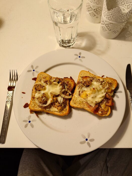 TvÄ rostade brödskivor med ost och kryddor pÄ en blommig tallrik, bredvid en gaffel och ett glas vatten pÄ ett bord.