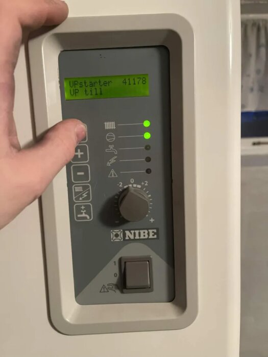 Inställningspanel för Nibe bergvärmepump med digital skärm och vred; visar texten "UPstarter 41178 UP till". En hand justerar panelen.