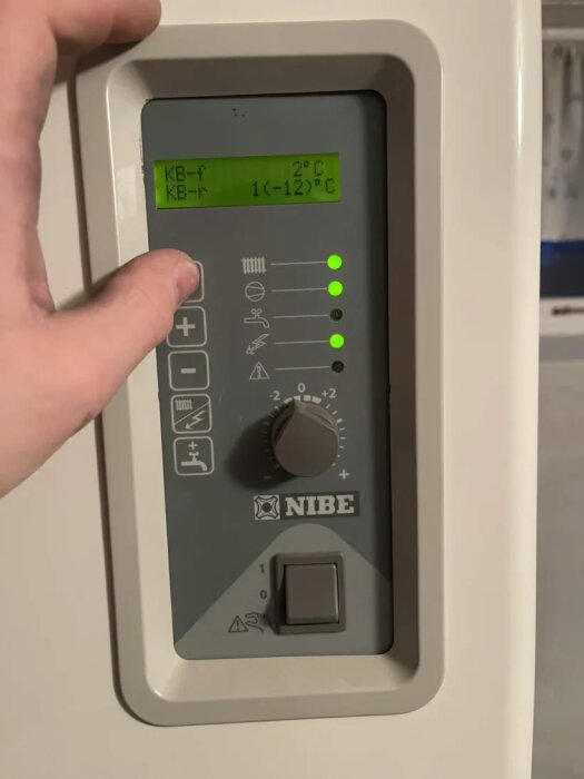 Nibe bergvärmepumpkontroll med grön digital display visar temperaturer och symboler. Vred står på 5.5 av 9. Hand justerar vredet.
