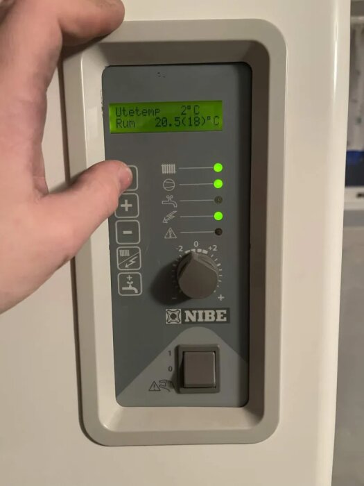 Nibe bergvärmepumps panel med temperaturinställning; utetemp 2°C, rumstemp 20.5°C. Hand justerar vredet. Lysande gröna indikatorlampor.
