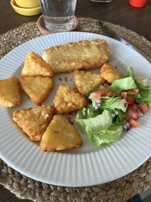 Torrt halvfabrikat med rösti, panerad torsk och fÀrsk sallad pÄ en vit tallrik med glas vatten i bakgrunden.