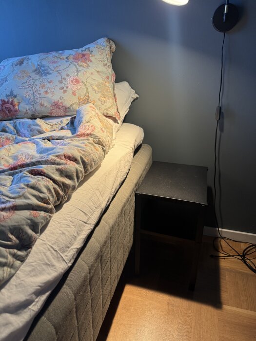 Säng med blommigt överkast och sängbord bredvid. Bordet är enkelt och svart, placerat på golvet längs en mörgrå vägg under en vägglampa.