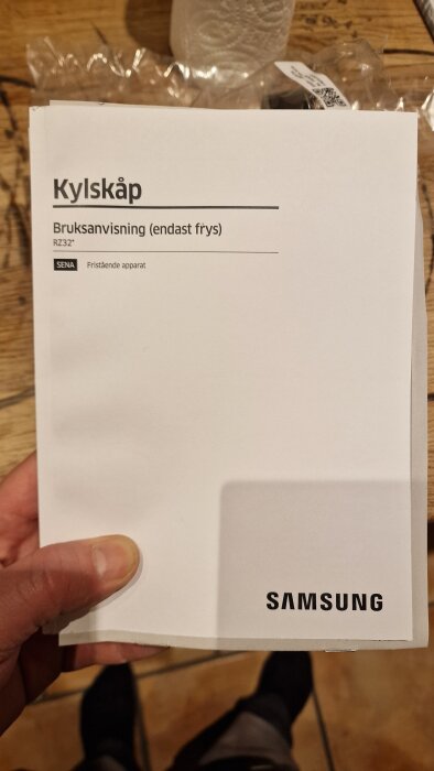 Bruksanvisning för Samsung-kylskåp (endast frys) hålls av en hand över ett träbord.
