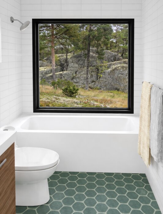 Modernt badrum med badkar och stort fönster som vetter mot skogslandskap. Vitt kakel och grönt hexagonkakel på golvet.