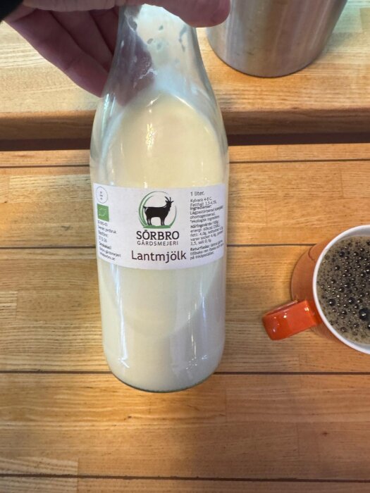 Flaska med ekologisk lantmjölk från Sörbro gårdsmejeri på ett träbord. Bredvid står en kopp med svart kaffe i en orange mugg.