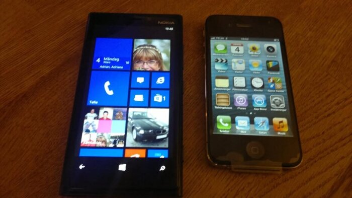 Två smartphones på ett bord, en med Windows Phone och en med iOS, visar startskärmar.
