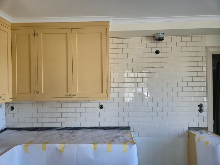 Gult köksskåp och kaklad vägg under renovering med skyddspapper på bänkskivan.