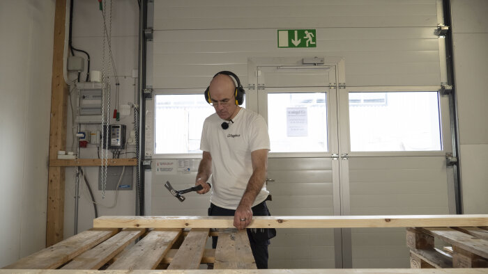 Man med hörselkåpor arbetar med en hammare vid en arbetsbänk i verkstadsmiljö.