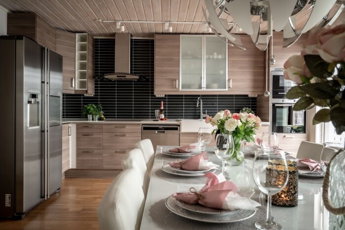 Modernt kök med svart kakel ovanför diskbänken, träskåp och rostfria vitvaror. Vitt dukat matbord med rosa servetter och blommor.