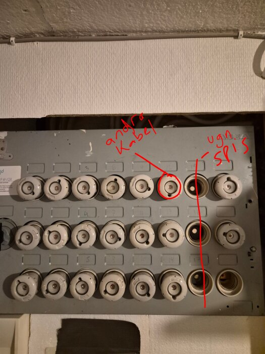 Äldre elcentral med porslinssäkringar, några saknas. Röd text och linjer pekar på specifika säkringar med märkning för byte av kabel.