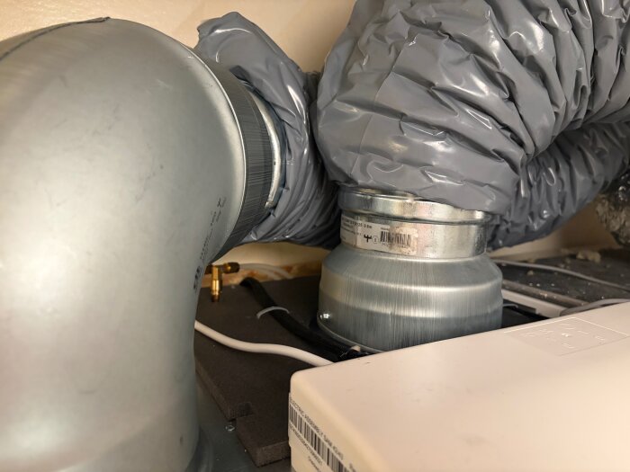 Grå ventilationsslang ansluten till ett metallrör i ett ventilationssystem, placerad nära en vit elektrisk enhet.
