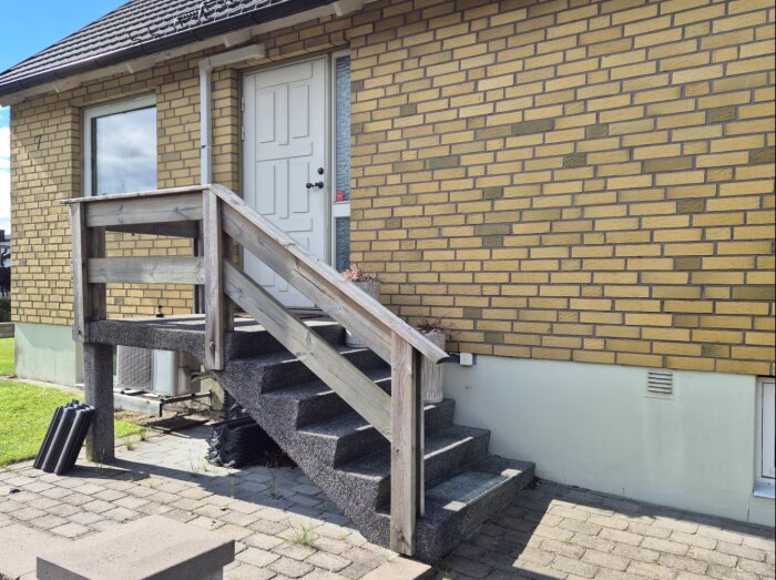 Ytterdörr med trappa och räcke i trä på ett tegelhus, omgiven av stenlagd gång och gräsmatta.