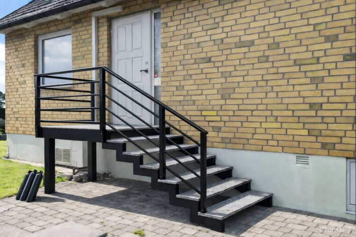 Modern stentrappa med svarta metallräcken leder upp till vit dörr på gul tegelfasad. Gräsmatta och stenlagd gång i förgrunden.