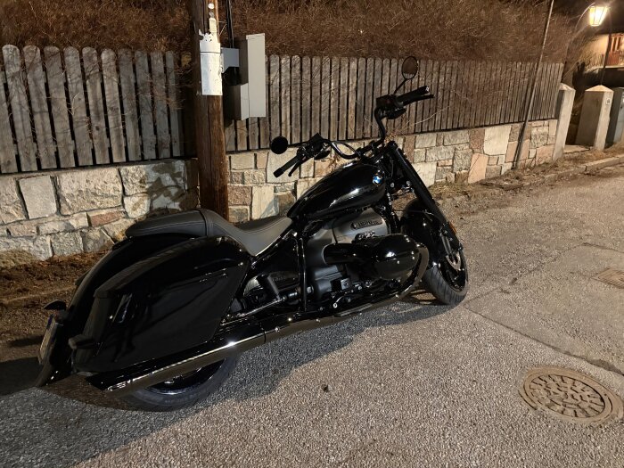 Svart motorcykel parkerad på en gata framför ett staket av trä och sten under natten, med gatubelysning som lyser upp området.