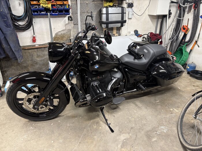 Svart motorcykel parkerad i ett garage med verktyg och utrustning på väggarna, skyddskläder och cykeldäck synliga i bakgrunden.