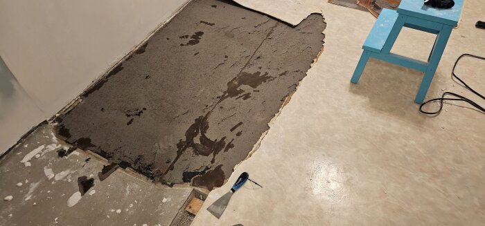 Rivet plastgolv i sovrum, synliga limrester och papp kvar på betonggolv med verktyg intill, blå pall bredvid.