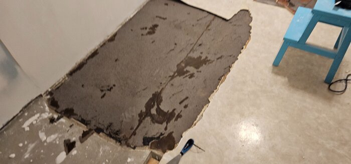 Golvet i ett sovrum där plastmattan delvis har rivits bort, visar underliggande papp och lim ovanpå betongplattan. En blå pall står intill.