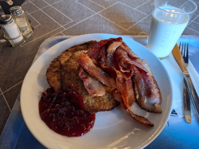 Raggmunk med stekt fläsk och lingonsylt på en tallrik, med ett glas mjölk bredvid på ett bordsklätt bord.