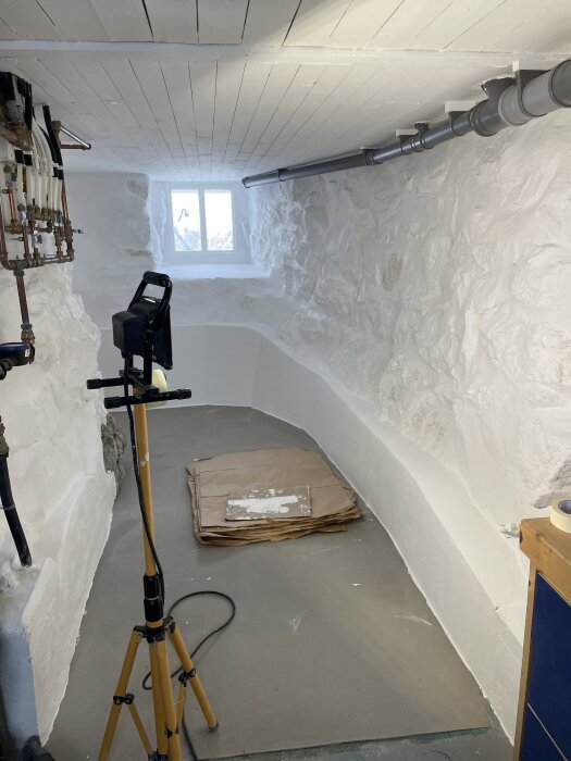 Källarutrymme under renovering, med vitmålade väggar och tak, synliga rör på väggen, ett litet fönster och en arbetsstrålkastare på stativ.
