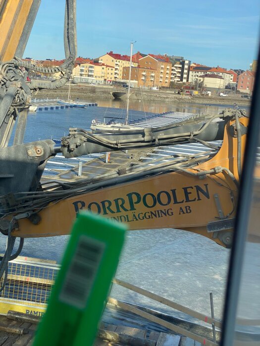 Grävmaskin vid vattnet med bostadshus i bakgrunden.