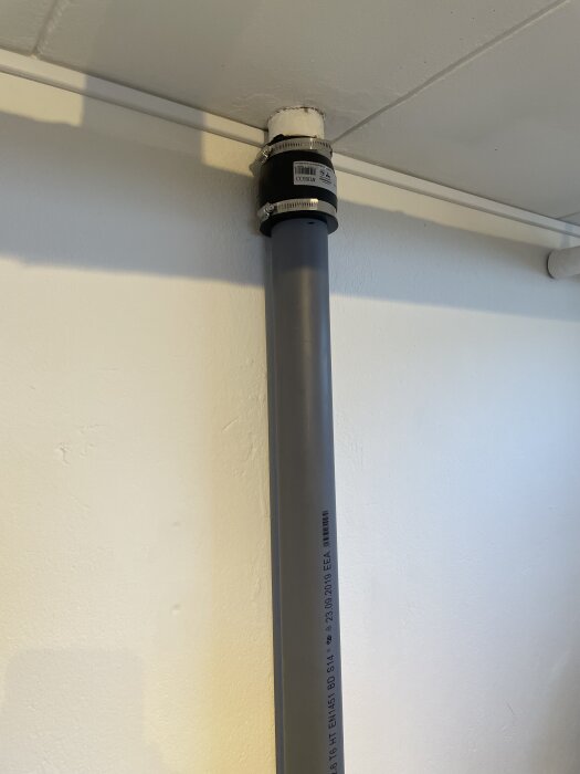 Grå avloppsrör går genom taket, anslutet med en svart gummimuff och metallklämmor, mot en ljus vägg.