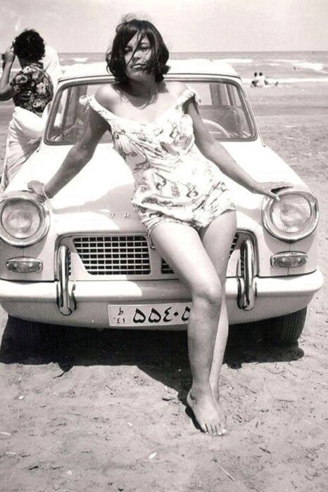 Kvinna i sommarklänning lutande mot en bil på en strand med havet i bakgrunden, vintage-stil från 1960-talet.