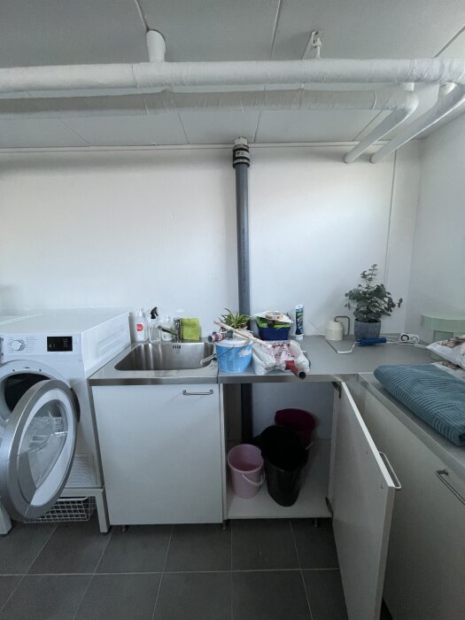 Grått avloppsrör går från tak till golv bakom skåp i tvättutrymme med vask, tvättmaskin, växter och rengöringsartiklar.