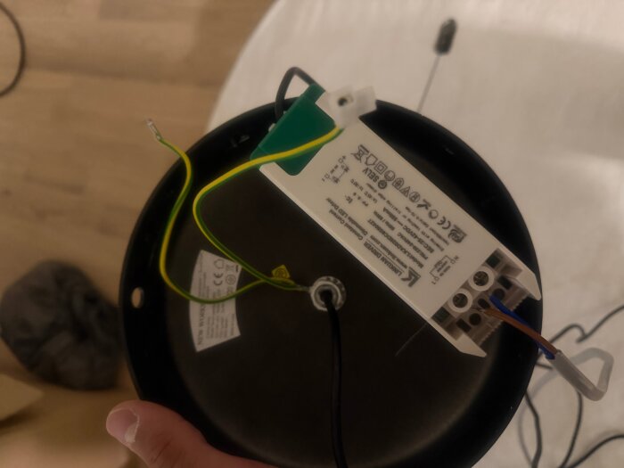 Elektronisk transformator monterad under en rund svart platta, exponerade elkablar anslutna, vit och grön färg på kontakter.