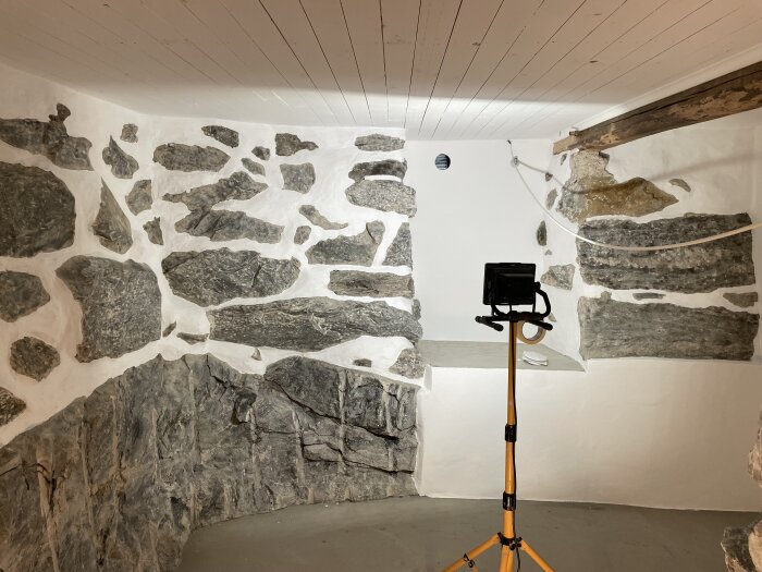 Stenklädd källarvägg med målad träpanel i taket och en arbetslampa på stativ i mitten av rummet.